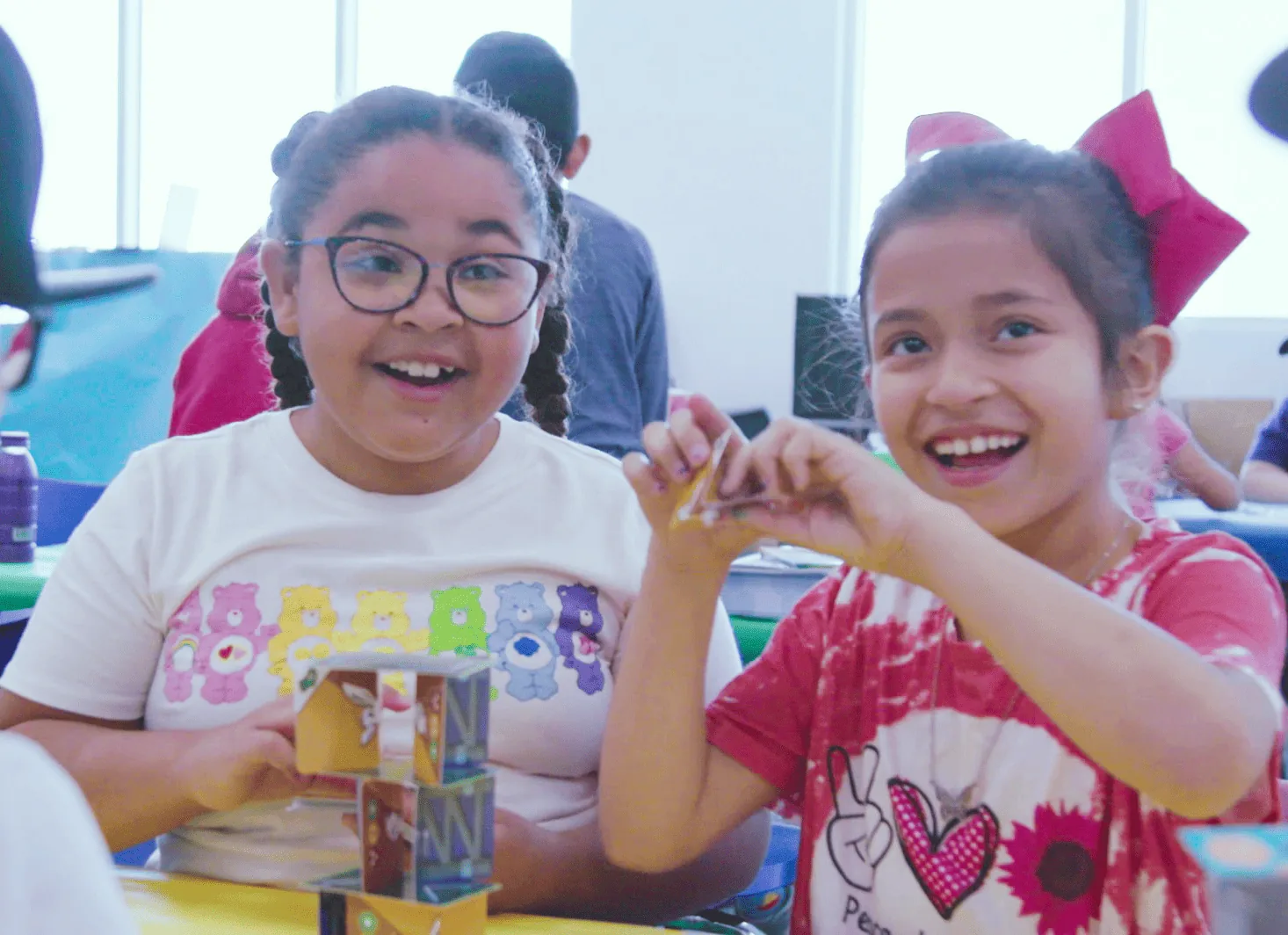 Two girls smile while building a small cube tower with craft materials at a table